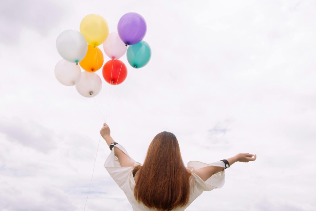 Young woman with arms wide open holds colorful balloons against a cloudy sky backdrop, embracing freedom and joy.